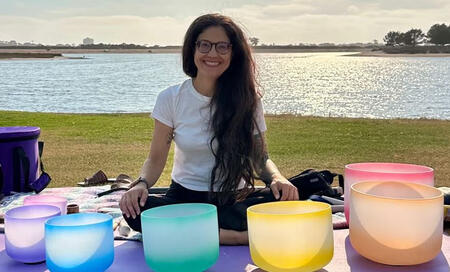 Sophia, founder of Sonic Lotus Yoga sitting in front of 7 rainbow colored chakra singing bowls with the Mission Bay water and grass behind her.